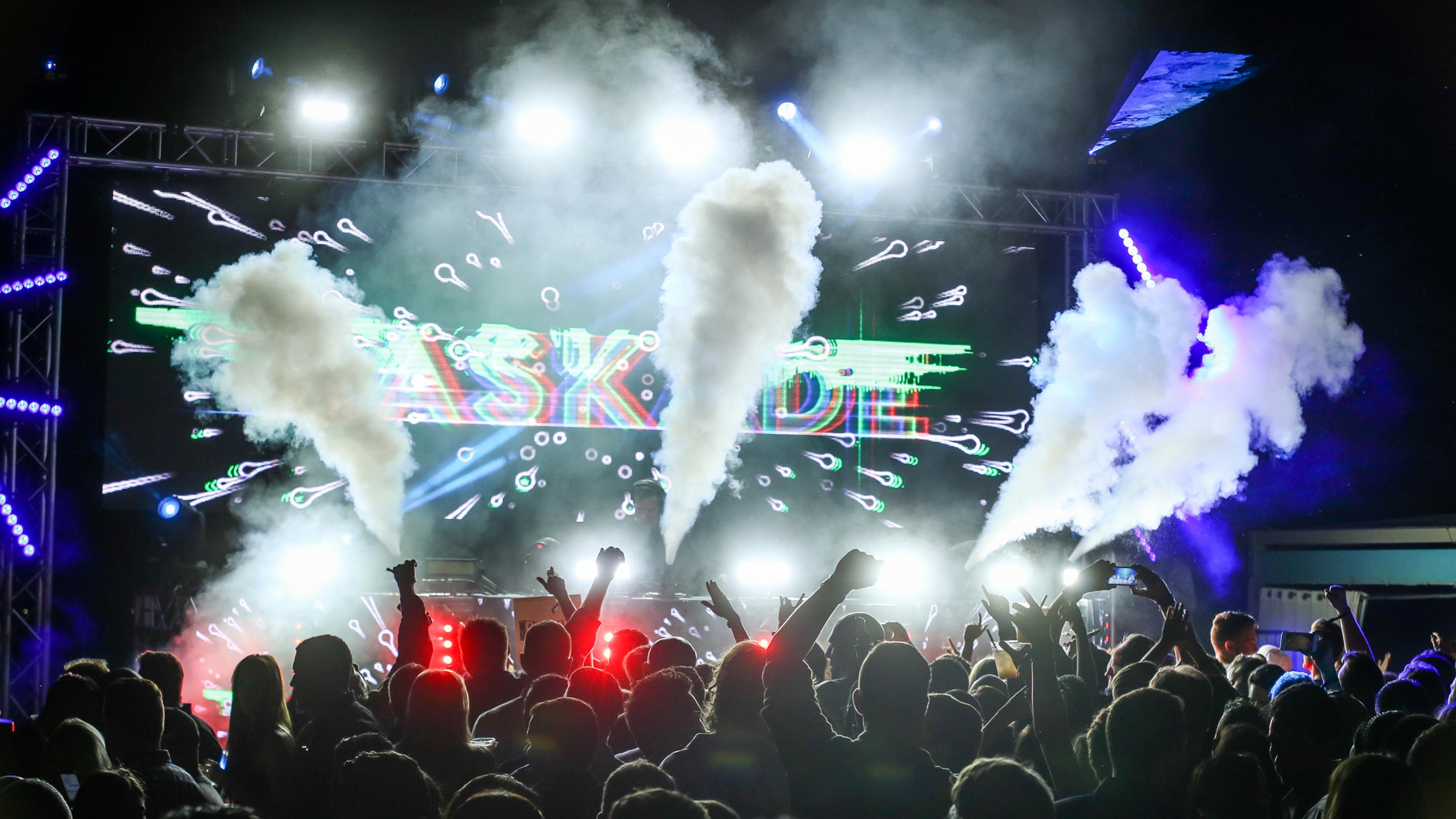 一位时尚的绿发女子坐在木楼梯上，身穿白色钩针连衣裙和黑色靴子，背景是一幅巨大的几何艺术品。