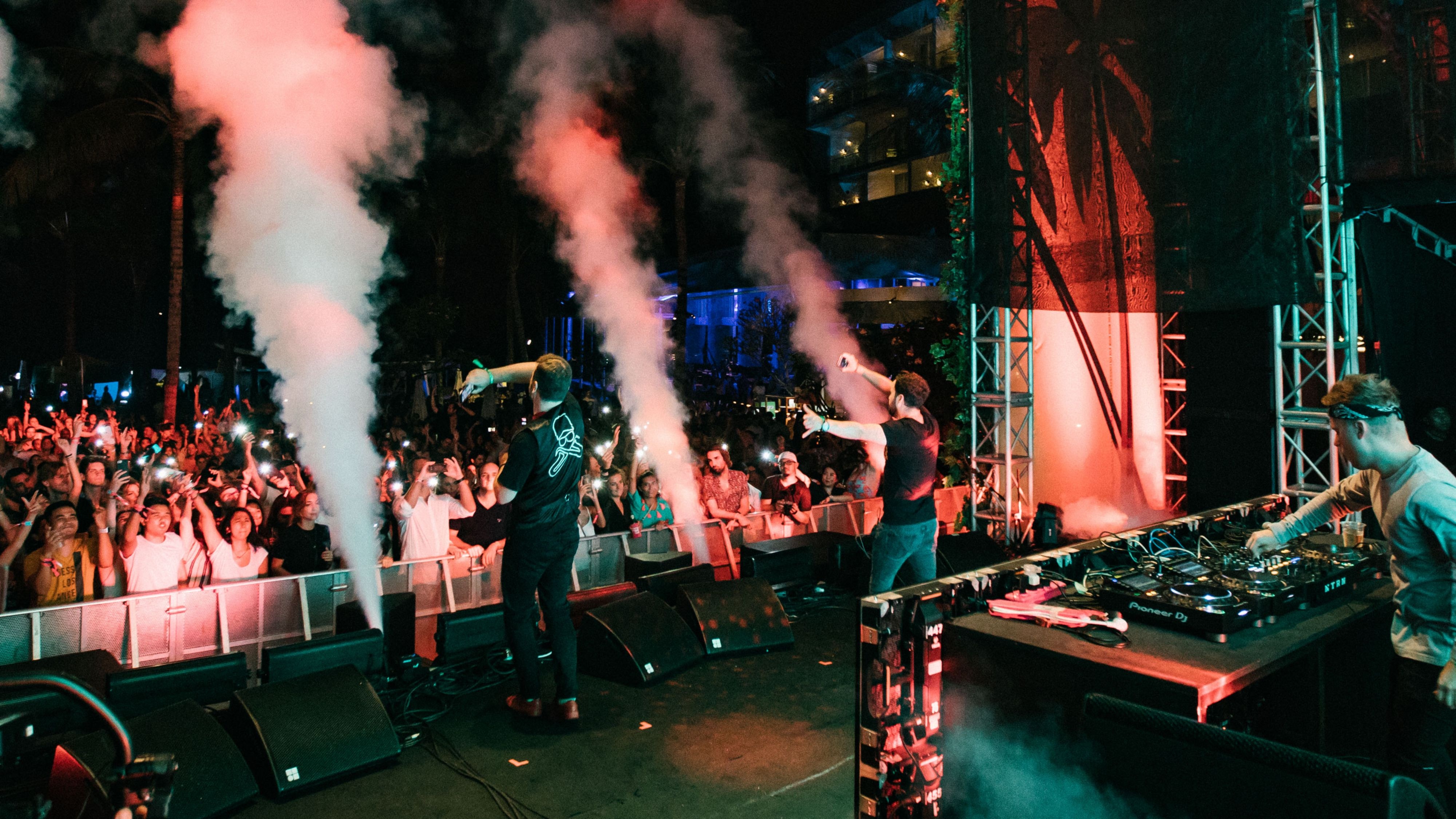 一位身穿扎染衬衫的 DJ 在俱乐部中溻音，后有摄影师团队在录影。​